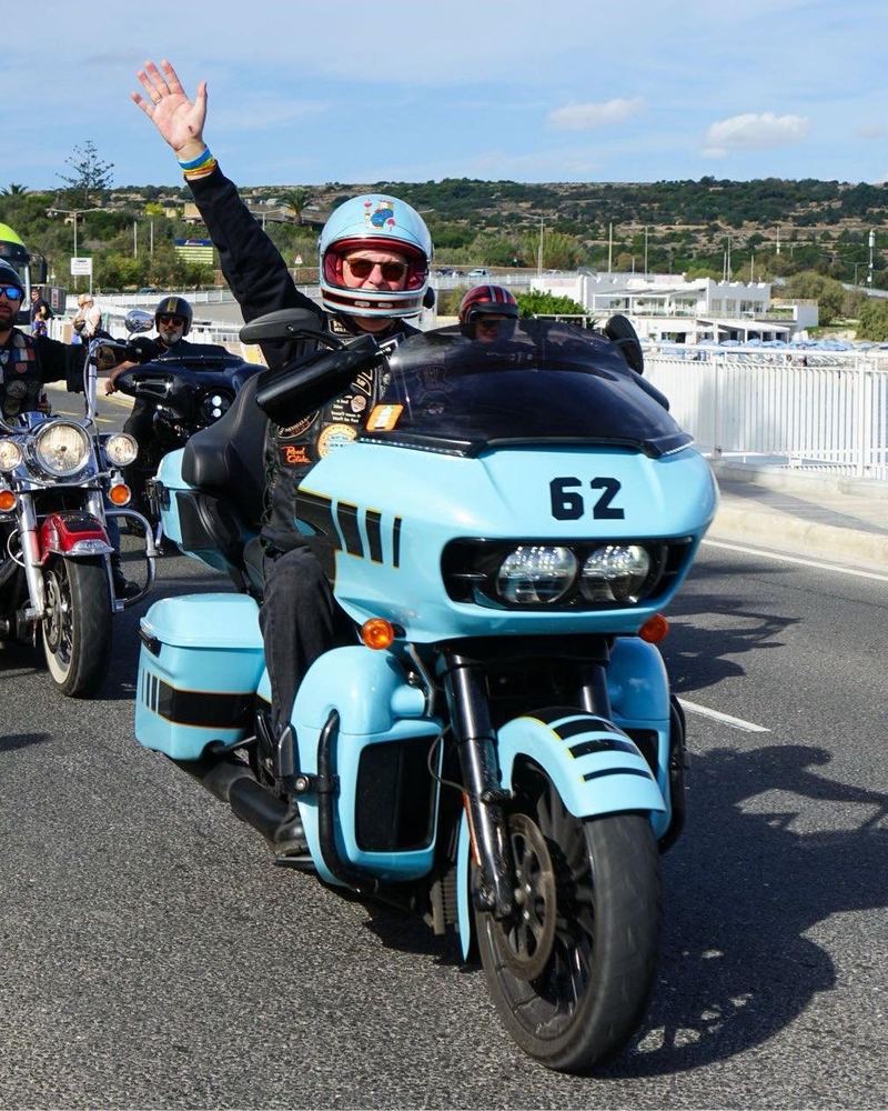 Person on a blue motorcycle with sidecar, waving, on a road with other motorcycles in the background.